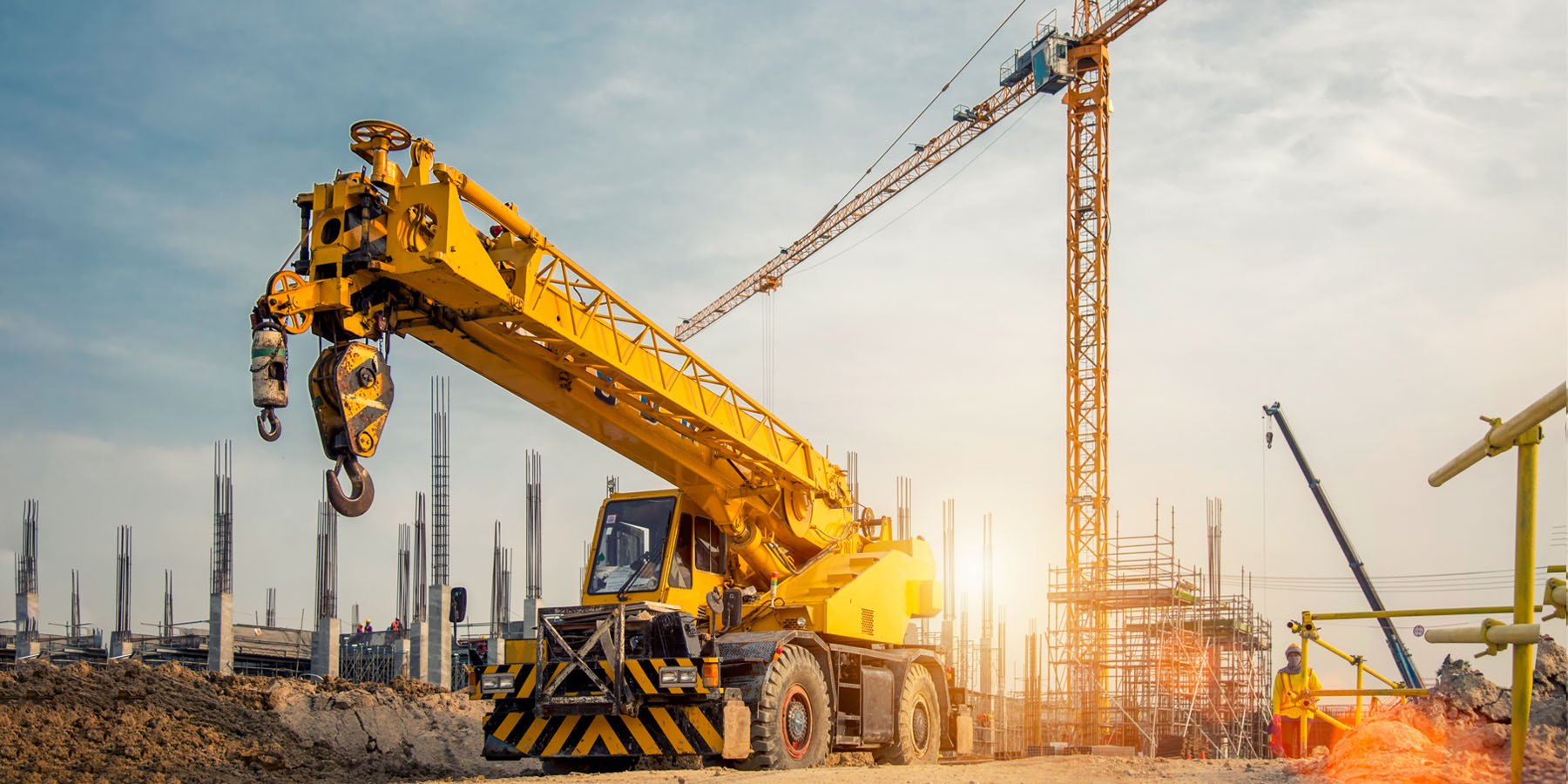 A rugged yellow mobile crane in front of a building construction site with a yellow tower crane in the background and workers on site in hi-visibility jackets